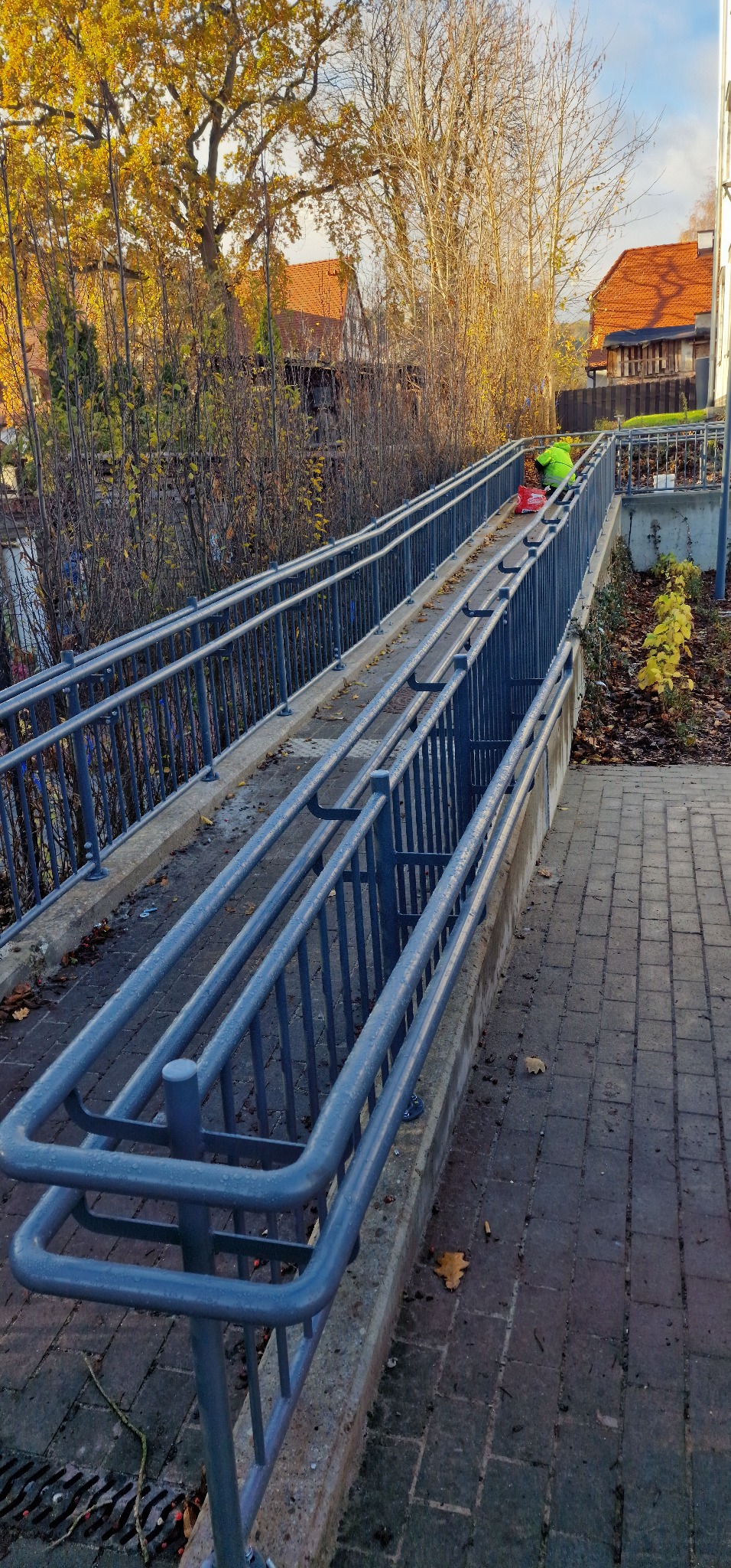 Szara metalowa balustrada na podjeździe z kostki brukowej, jesienne otoczenie, osoba w zielonej kurtce w tle. Inwestycja w Sopocie.