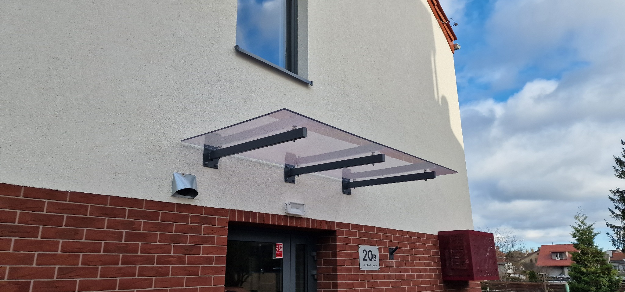 Szklany daszek nad wejściem do budynku z ceglaną elewacją i numerem 20b. Widoczne niebo i fragment drzewa. Inwestycja w Sopocie.