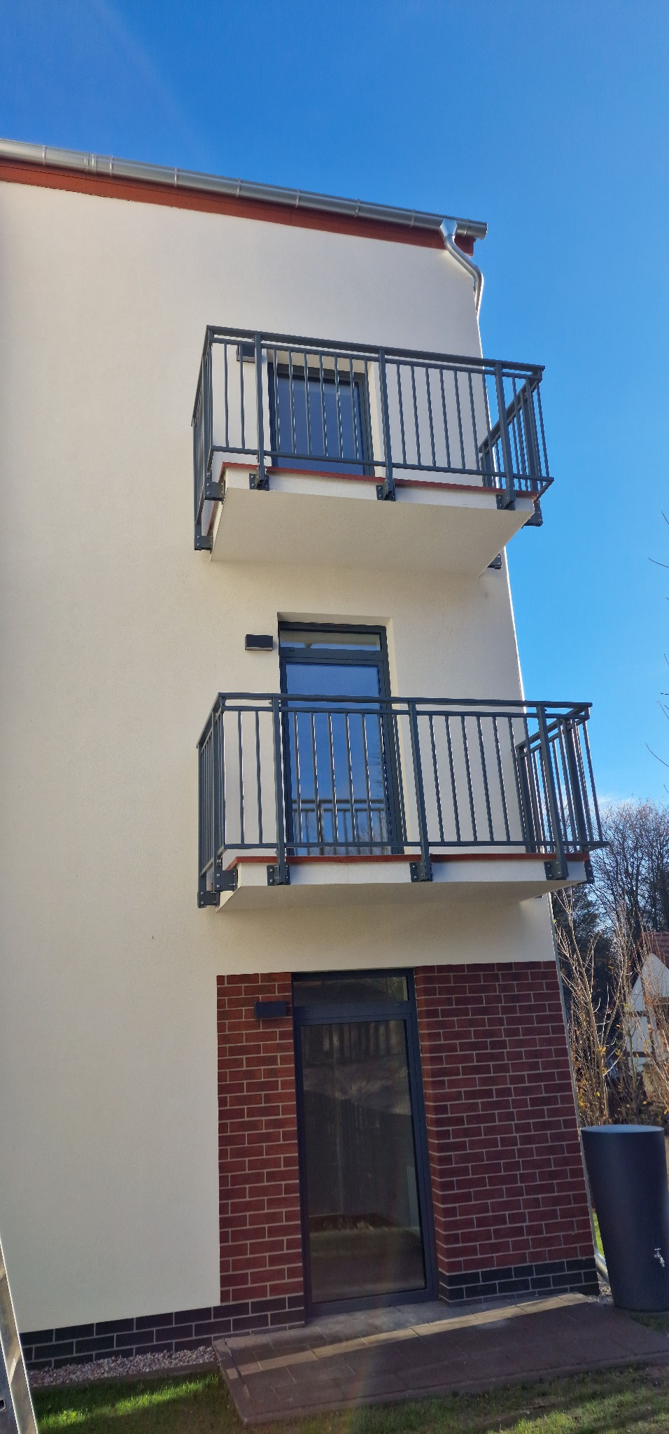 Elewacja budynku z dwoma balkonami z szarymi, metalowymi balustradami. Widok z dołu, błękitne niebo w tle. Inwestycja w Sopocie.