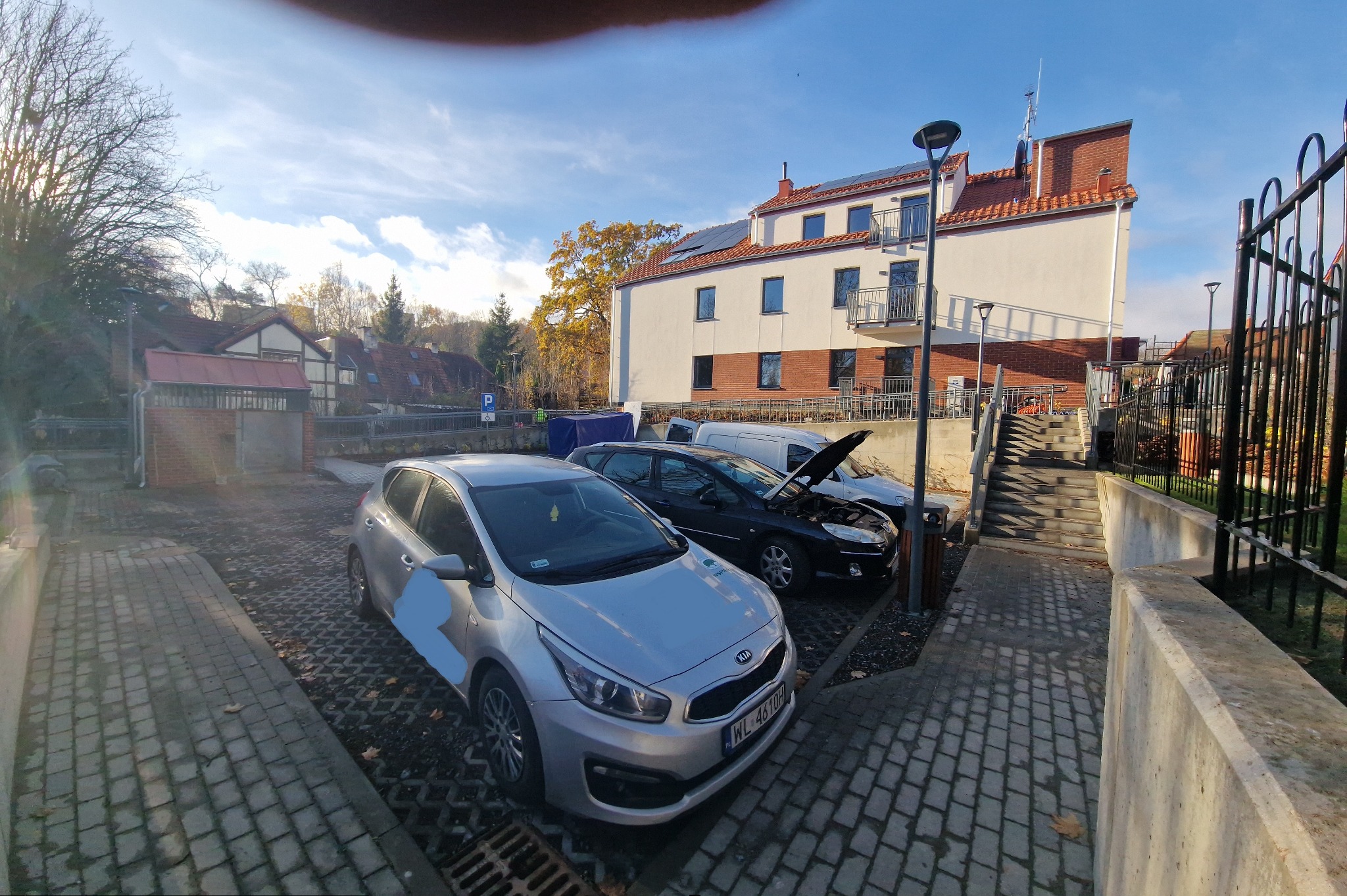 Metalowe ogrodzenie posesji w Warszawie, widoczne zza zaparkowanych samochodów. W tle budynek z balkonami i panelami słonecznymi na dachu.