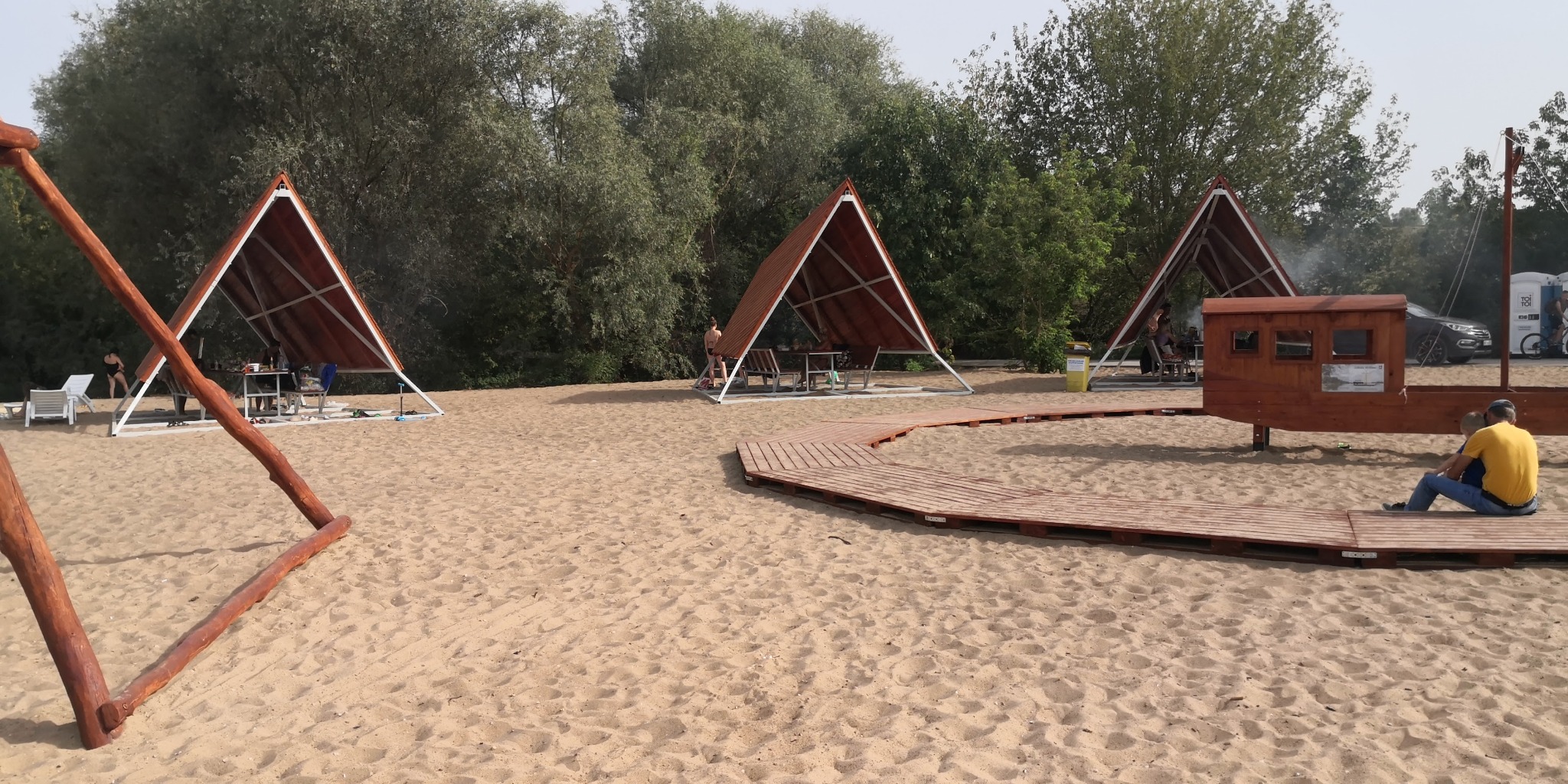 Plaża z drewnianymi altanami o trójkątnej konstrukcji, połączonymi ścieżką z desek. W tle zieleń drzew i krzewów. Elementy drewniane, piasek, letni dzień.