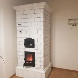 Biały piec kaflowy Kuzniecowa z czarnymi drzwiczkami paleniska i piekarnika, widoczne płomienie wewnątrz, minimalistyczne wnętrze z elementami instalacji na ścianie.