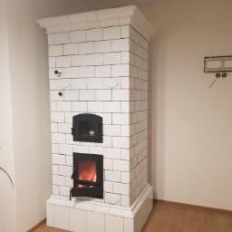 Biały piec kaflowy Kuzniecowa z czarnymi drzwiczkami paleniska i piekarnika, widoczne płomienie wewnątrz, minimalistyczne wnętrze z elementami instalacji na ścianie.