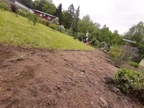 Przygotowanie terenu pod trawnik na nierównym terenie, widoczna przekopana ziemia, częściowo zielona trawa, w tle dom z czerwonym dachem i pracujące osoby.