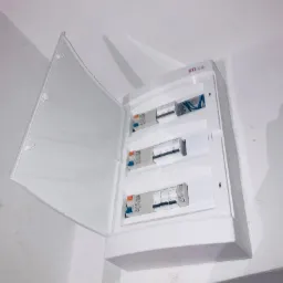 Otwarta, biała skrzynka elektryczna z bezpiecznikami Schneider Electric zamontowana na białej ścianie, widoczne przewody w górnej części.