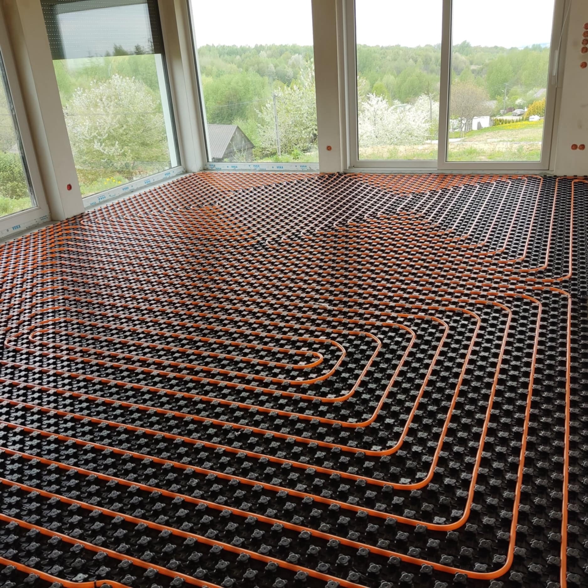 Pomarańczowe rury systemu ogrzewania podłogowego ułożone na czarnej macie izolacyjnej w jasnym pomieszczeniu z dużymi oknami z widokiem na zielony krajobraz.