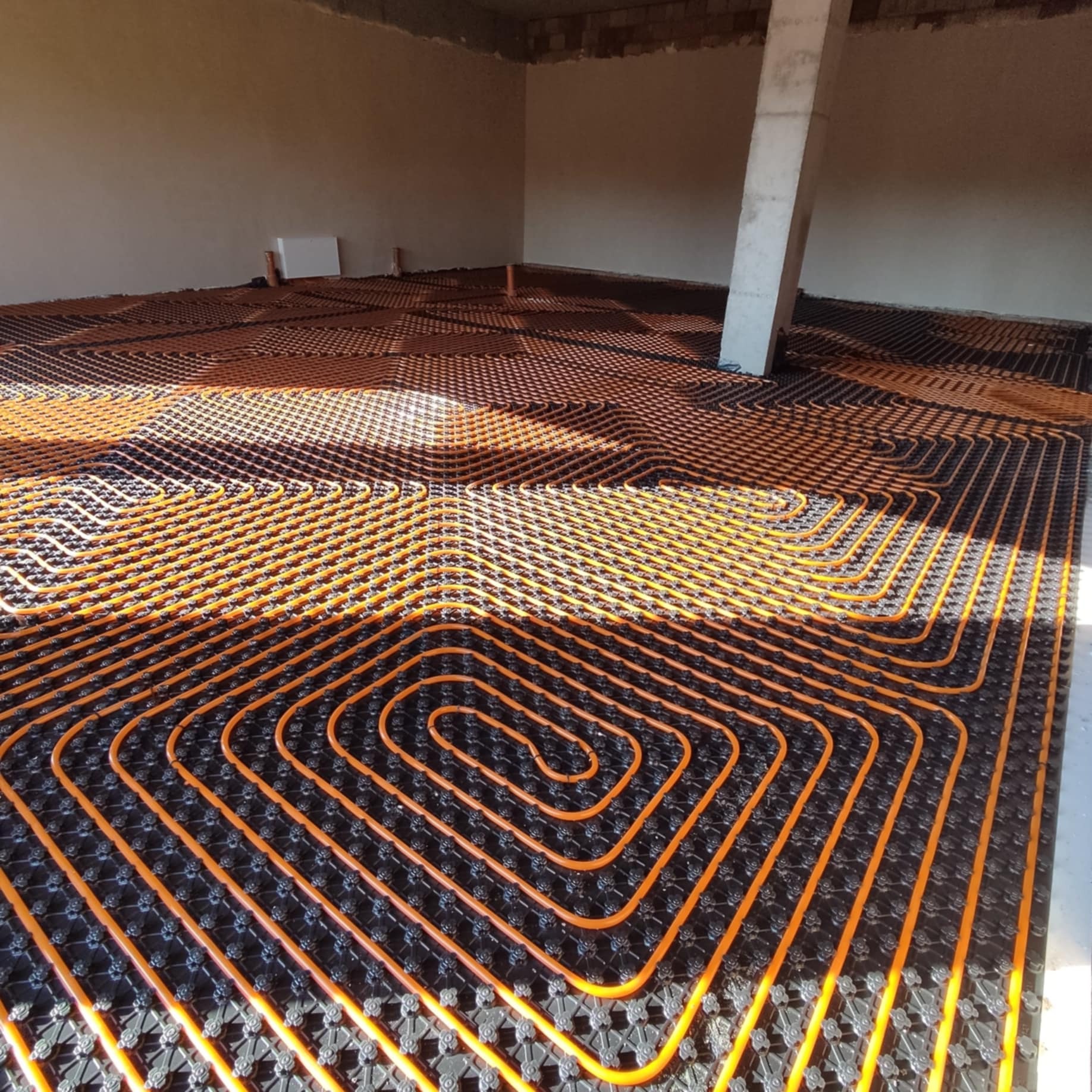 Pomarańczowe rurki grzewcze ułożone spiralnie na czarnej macie montażowej, przygotowane do zalania wylewką w surowym pomieszczeniu z betonowym słupem.