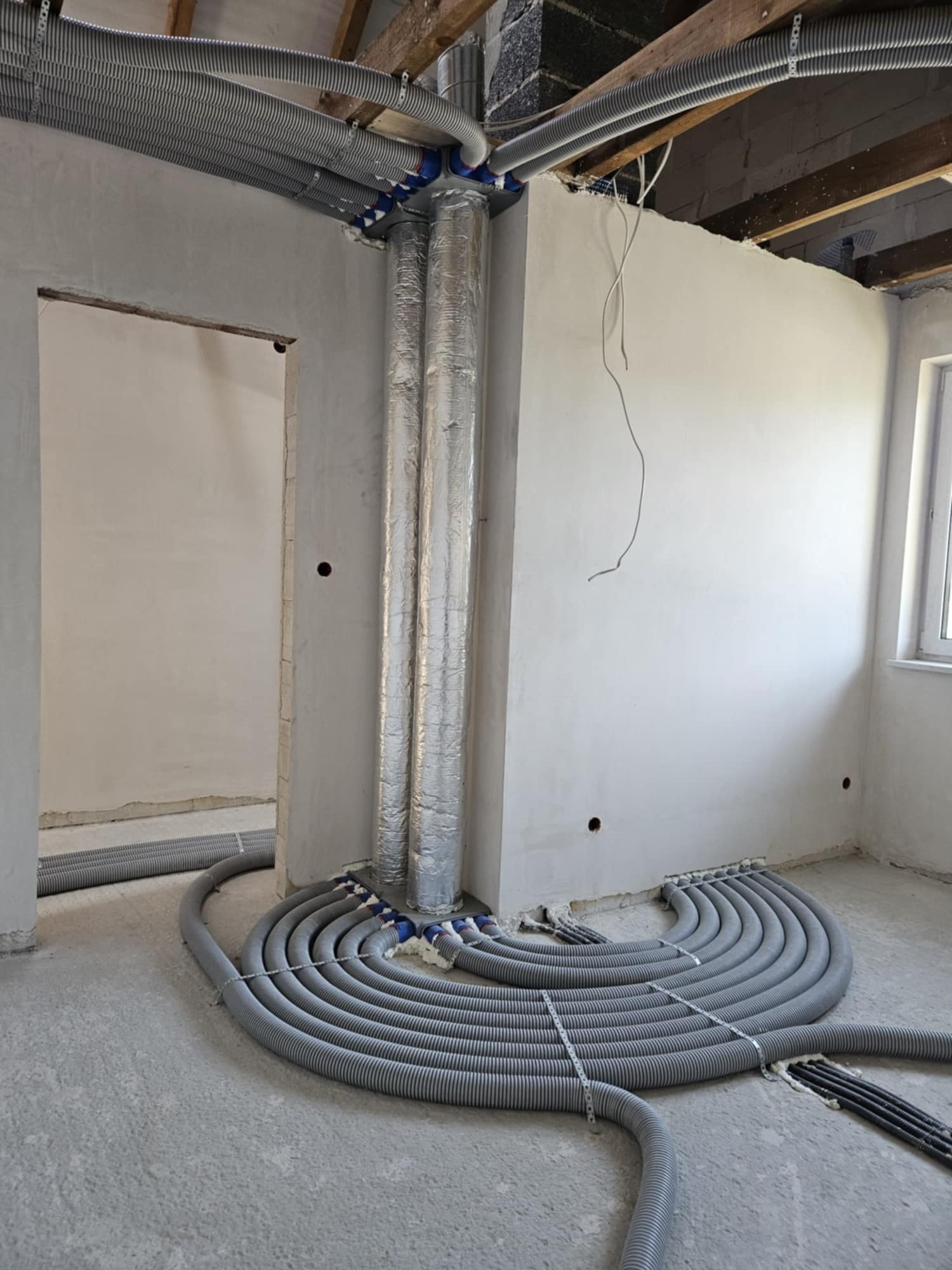 Szare, karbowane rury wentylacyjne ułożone spiralnie na surowym betonowym podłożu w pomieszczeniu w trakcie budowy, z widocznymi przewodami pionowymi owiniętymi folią aluminiową i drewnianą...