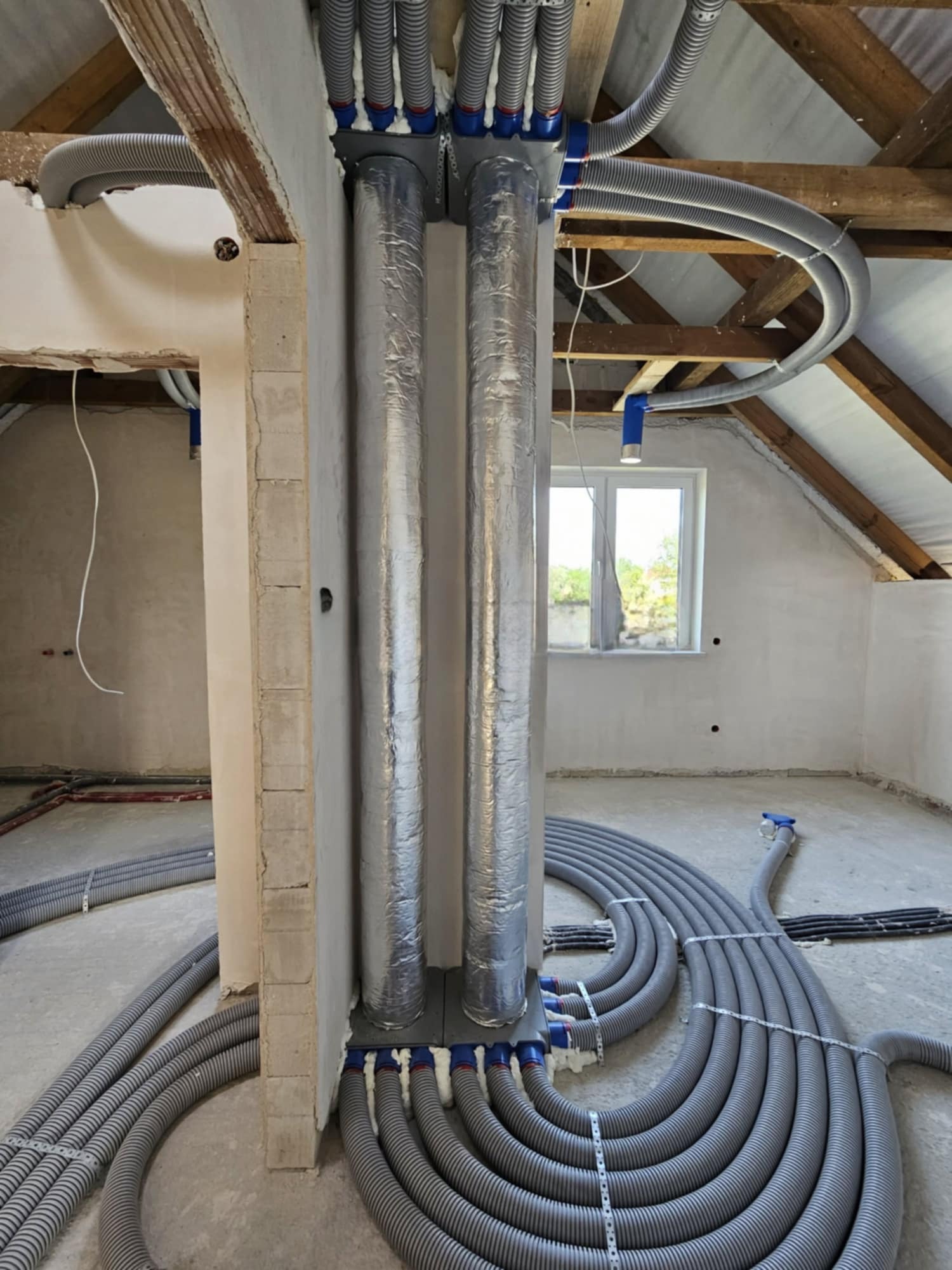Instalacja rekuperacji w surowym stanie poddasza: pionowe, owinięte folią przewody wentylacyjne oraz liczne elastyczne rury rozprowadzające, mocowane do stropu z drewnianymi belkami.