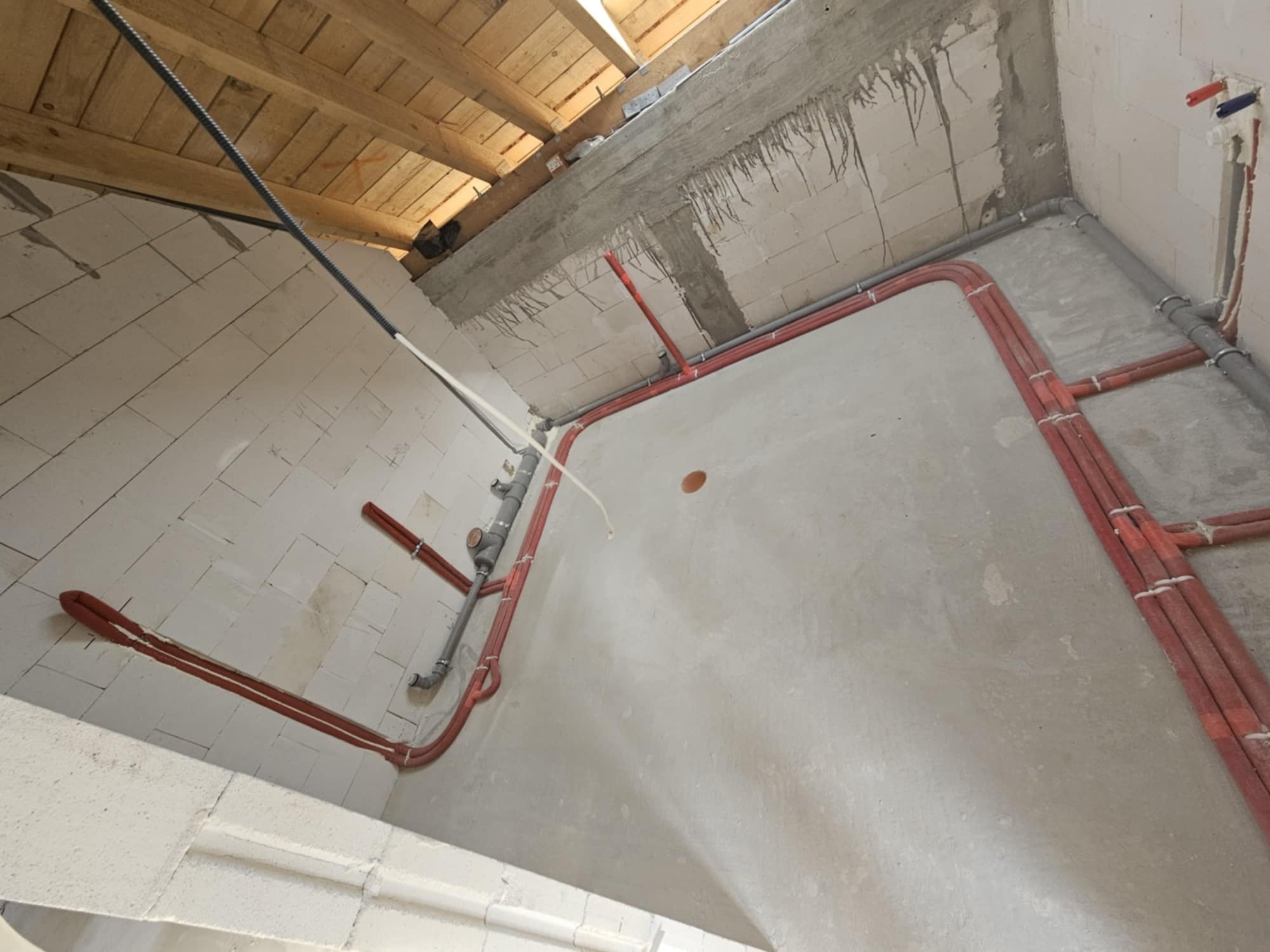 Surowy stan wnętrza z widocznymi instalacjami: rury grzewcze w izolacji i szare rury kanalizacyjne na ścianach z betonu komórkowego i poddaszu z drewnianych belek.