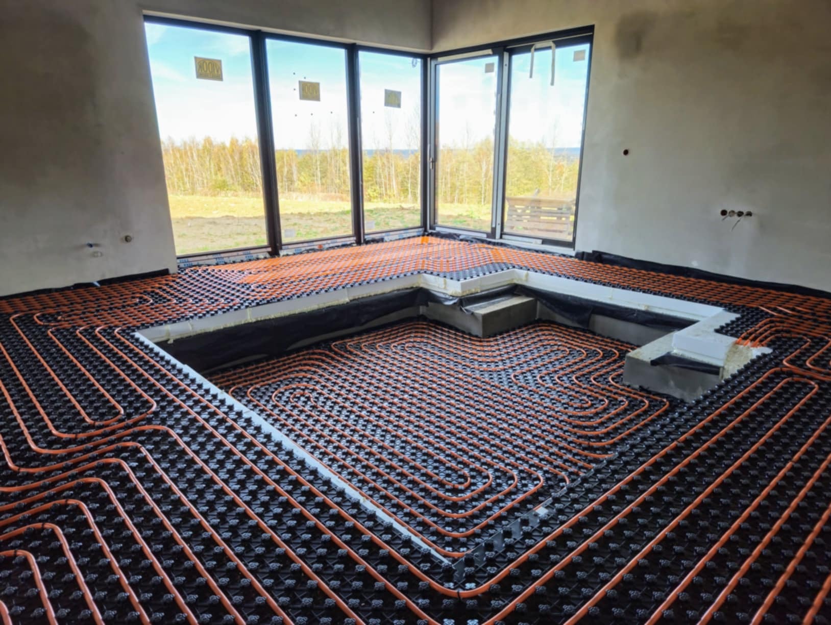 Pomarańczowe rurki instalacji grzewczej ułożone w pętle na czarnej macie z wypustkami, widoczne fragmenty izolacji i surowy beton ścian w nowym budynku z dużymi oknami.