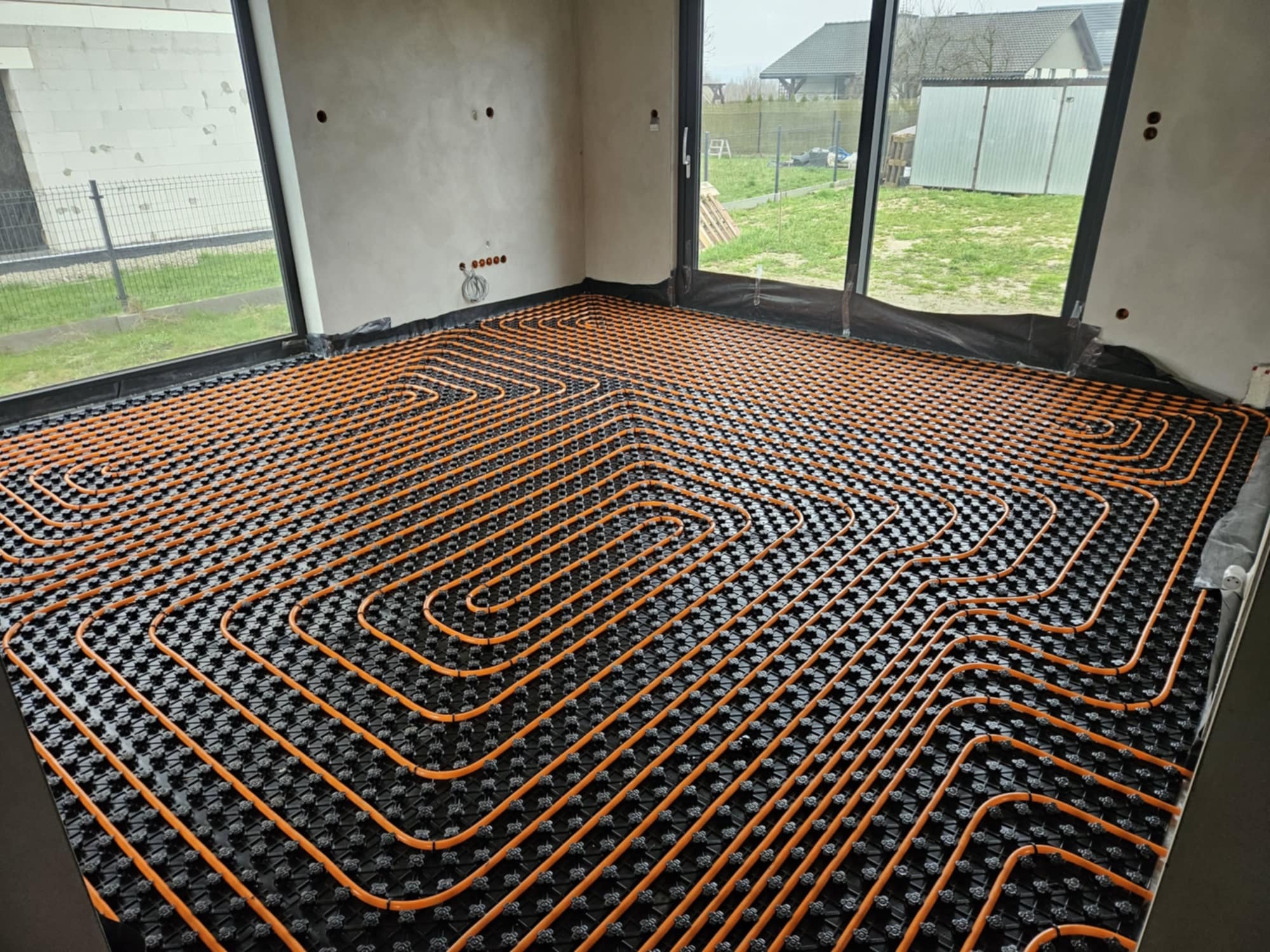 Pomarańczowe rurki instalacji grzewczej ułożone na czarnej macie w surowym pomieszczeniu z dużymi oknami, widok na zewnątrz.