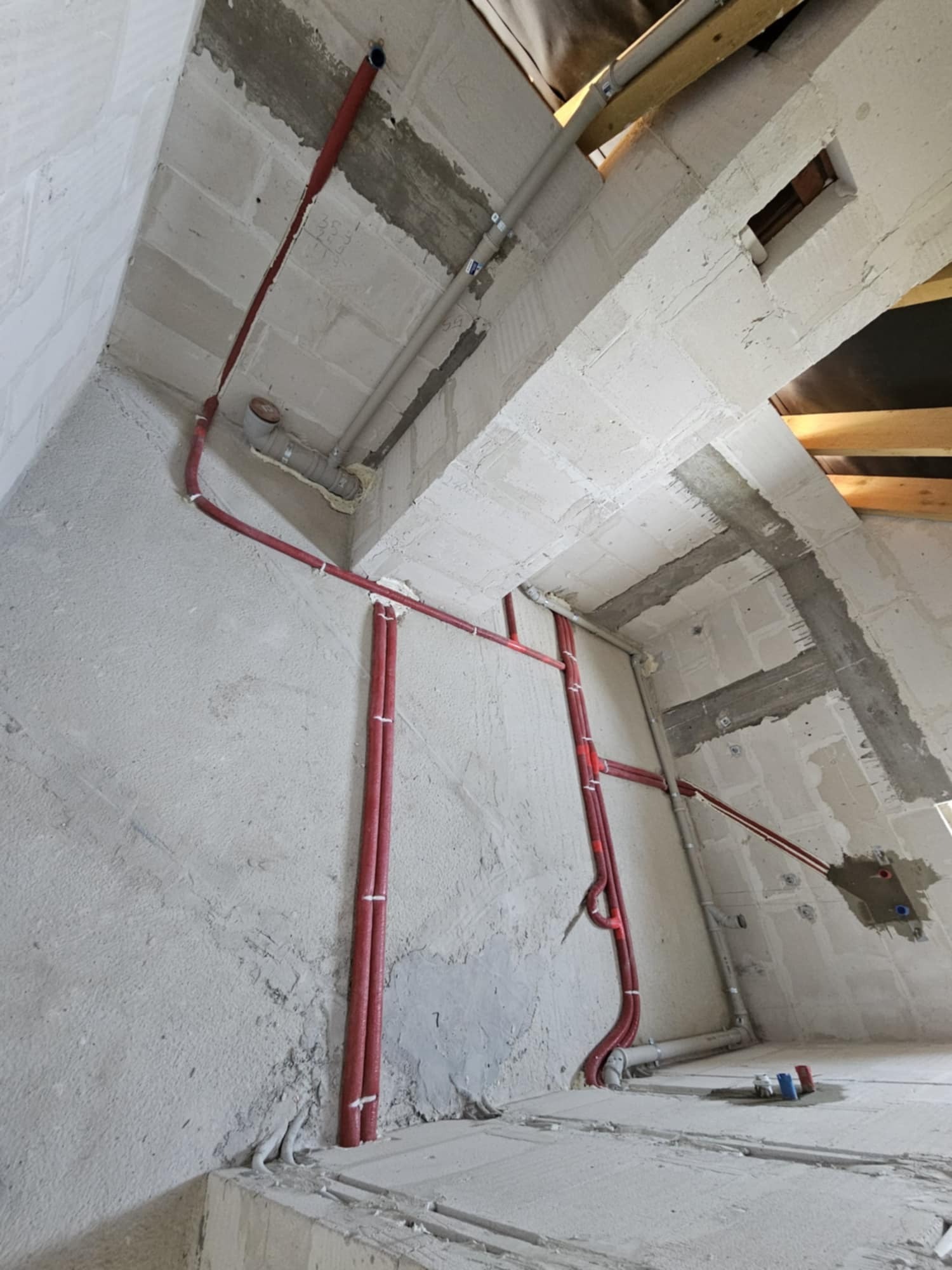 Nowe instalacje rurowe w surowym wnętrzu poddasza, widoczne czerwone rury grzewcze i szare rury kanalizacyjne, zamocowane do ścian z betonu komórkowego.