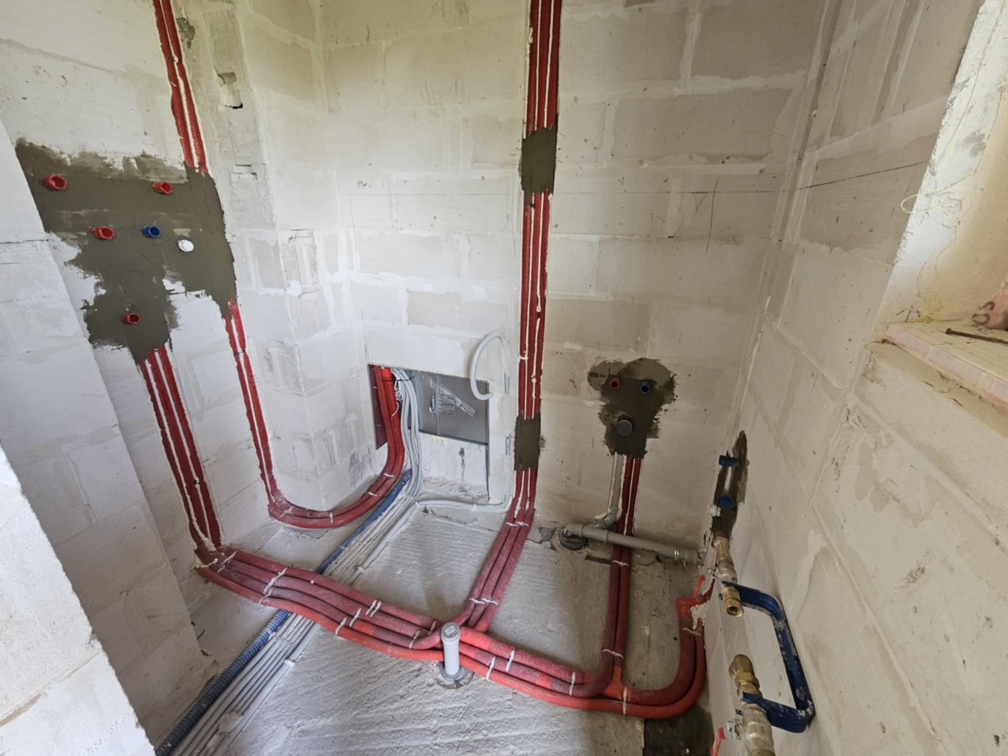 Surowe ściany z betonu komórkowego z rozprowadzonymi rurami w izolacji koloru czerwonego, częściowo zamurowanymi, oraz niebieskimi rurami instalacji elektrycznej, widoczne przyłącza wodne...