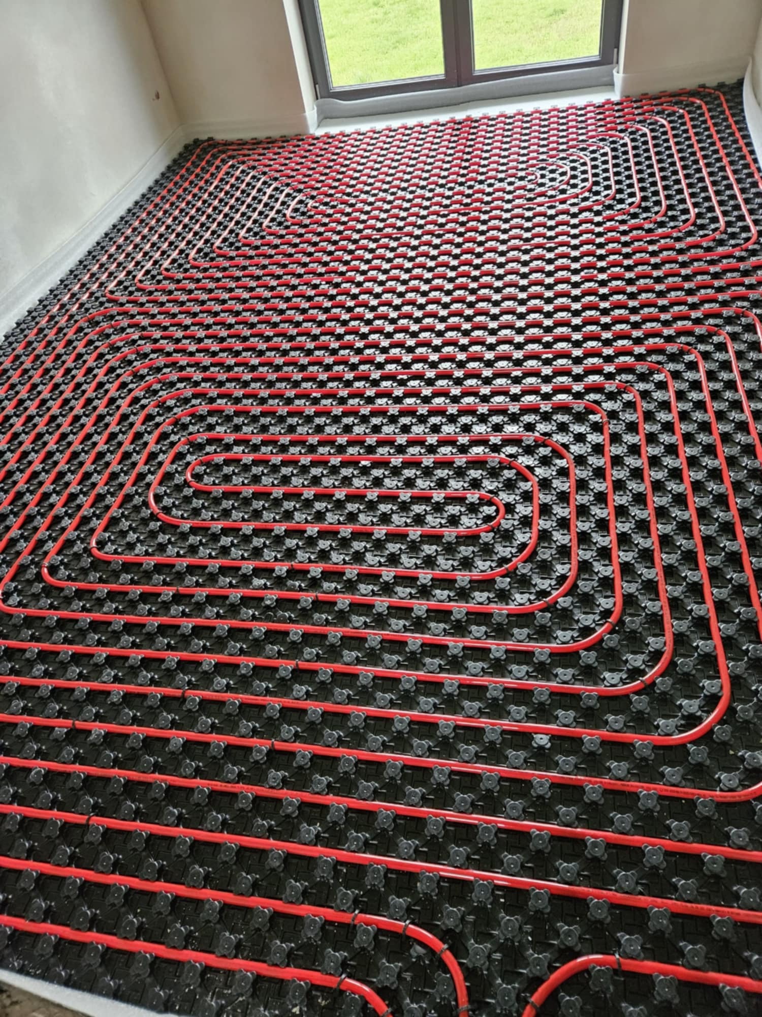 Instalacja ogrzewania podłogowego z czerwonymi rurami ułożonymi na czarnej macie montażowej, widok z góry na całą powierzchnię podłogi w jasnym pomieszczeniu z oknem.