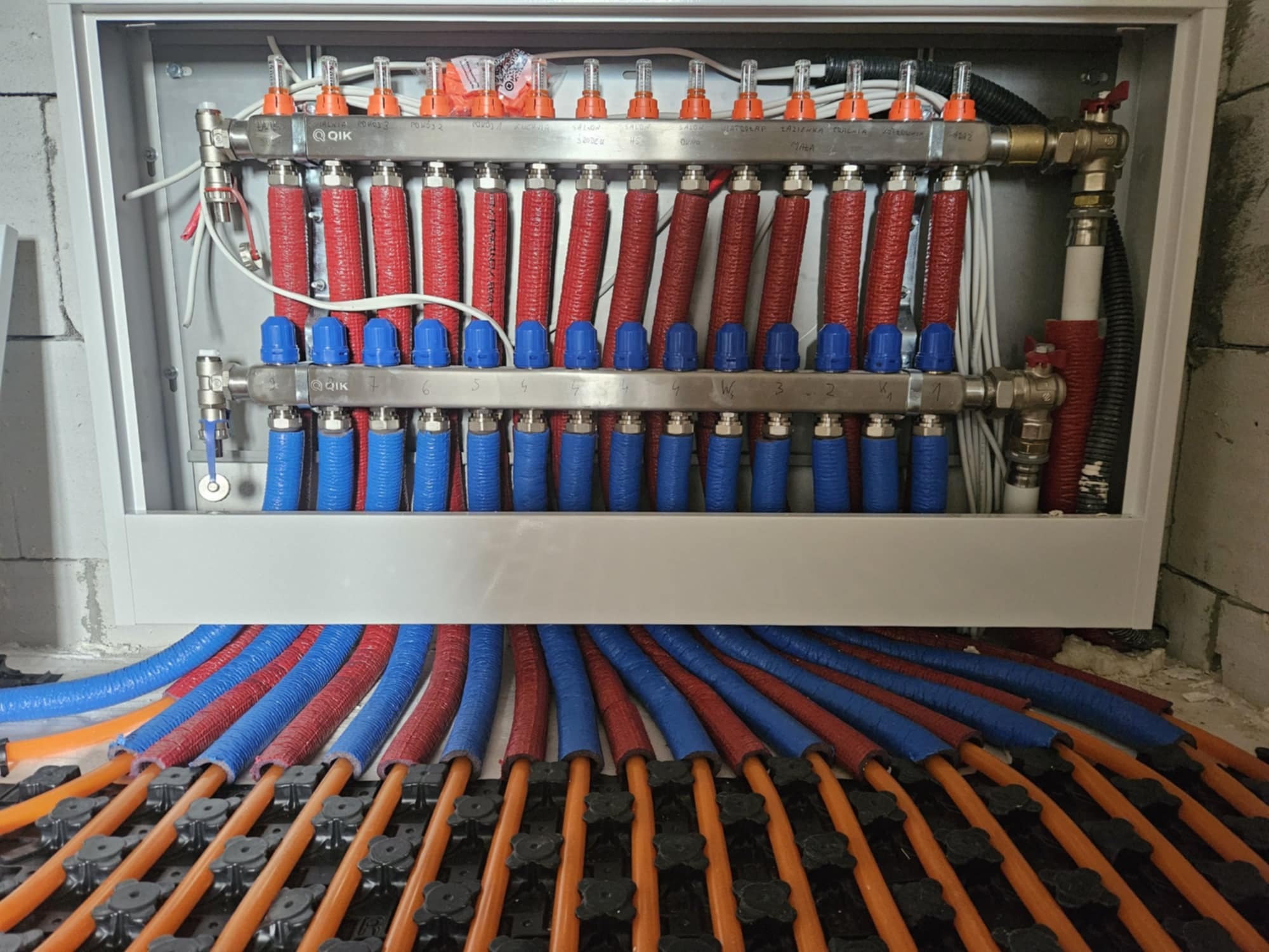 Rozdzielacz instalacji grzewczej podłogowej z rurami w izolacji czerwonej i niebieskiej, ułożonymi na czarnej macie montażowej z pomarańczowymi rurkami, widok z góry na instalację w trakcie montażu.