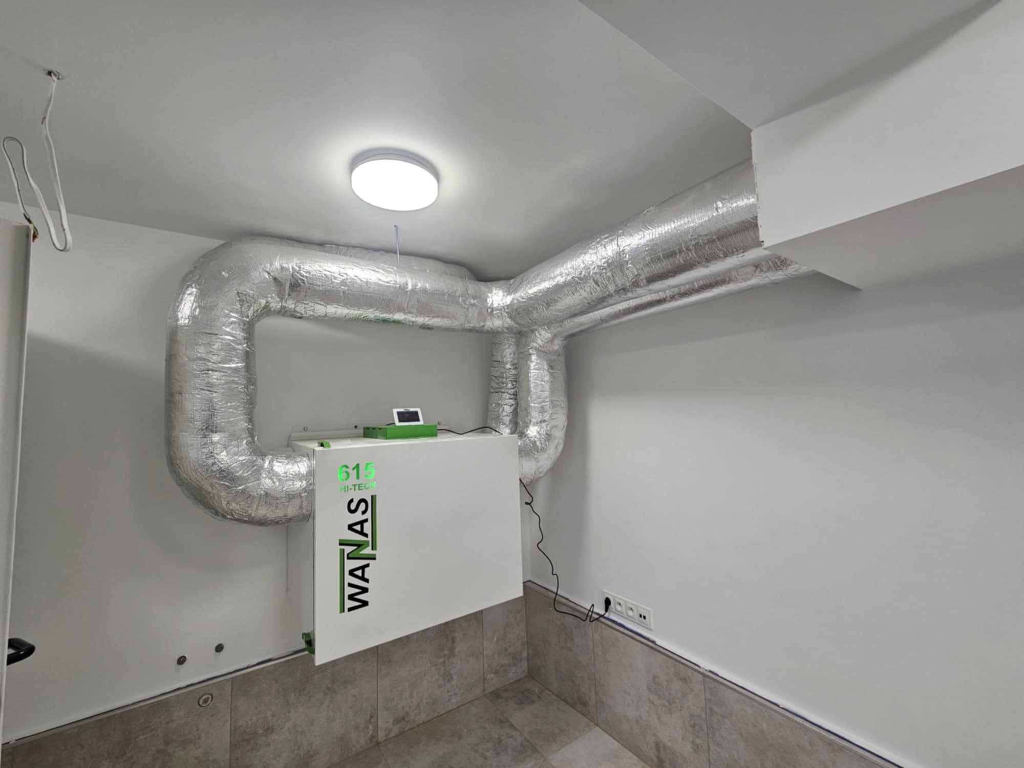 Centrala wentylacyjna z rekuperacją firmy WANAS, model 615 HI-TEC, z podłączonymi kanałami wentylacyjnymi w pomieszczeniu z szarą podłogą i białymi ścianami.