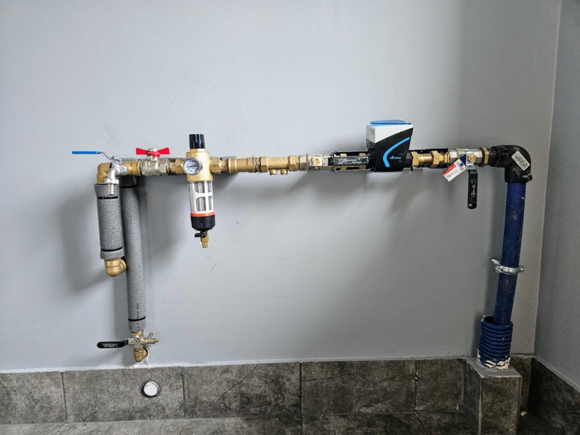 Fragment instalacji wodnej z filtrem, zaworami i licznikiem, przy szarej ścianie i ciemnej podłodze wyłożonej płytkami.