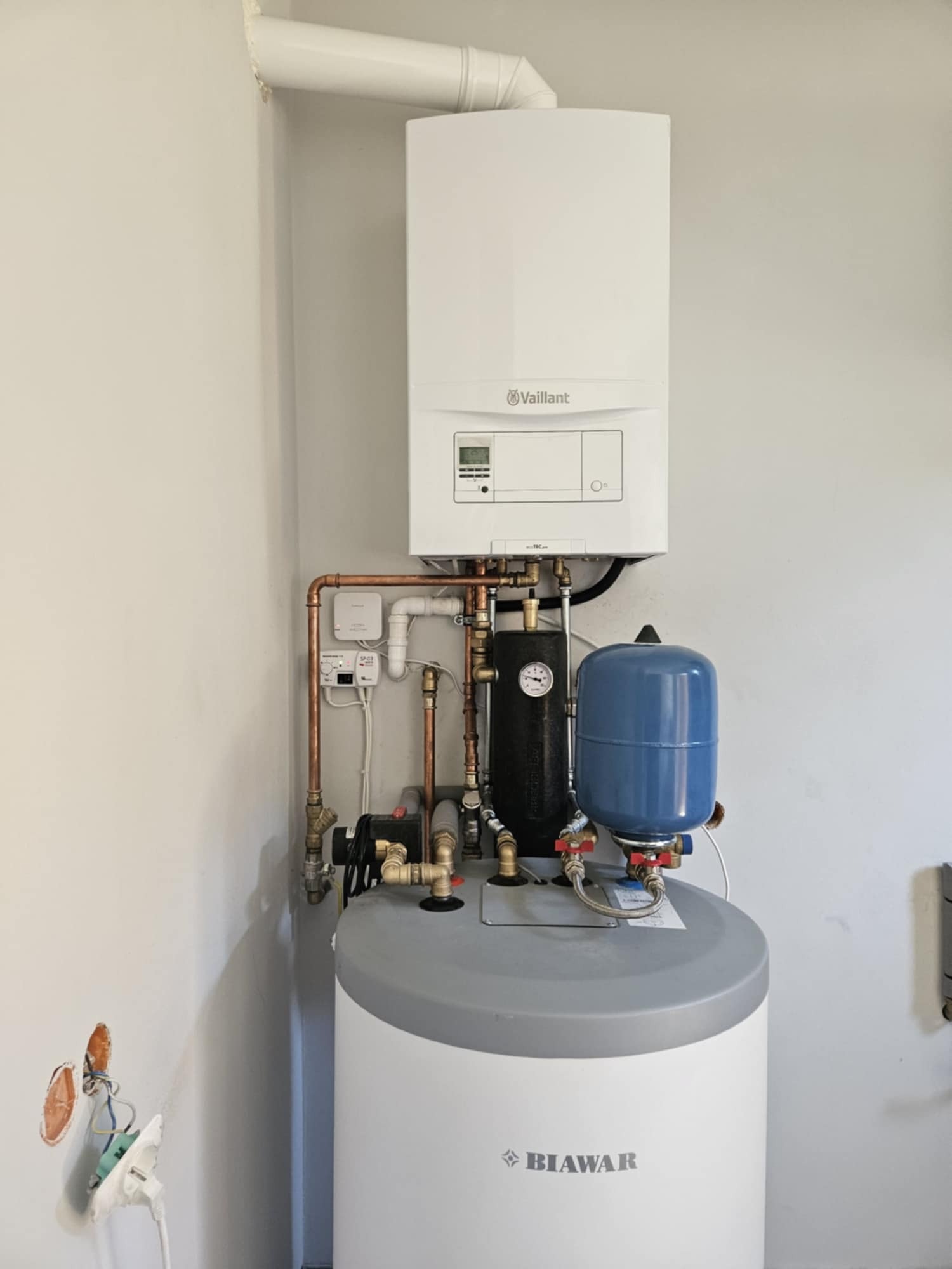 Kocioł gazowy Vaillant zamontowany nad biało-szarym zasobnikiem Biawar, połączony miedzianymi rurami i elementami hydraulicznymi, z widocznymi przewodami w ścianie obok.