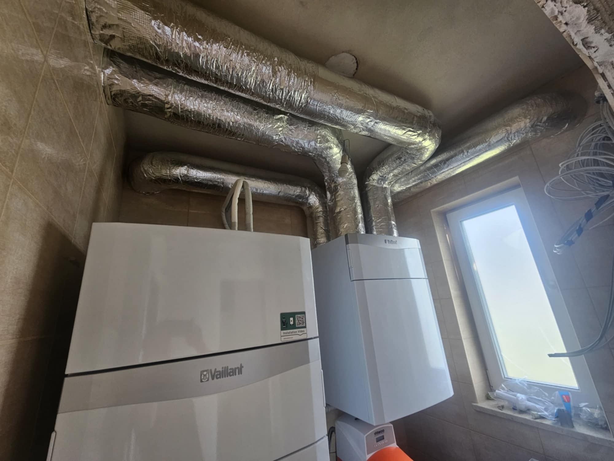 Dwa kotły grzewcze marki Vaillant zamontowane w pomieszczeniu z rurami wentylacyjnymi owiniętymi folią aluminiową, widoczne okno z akcesoriami hydraulicznymi na parapecie.