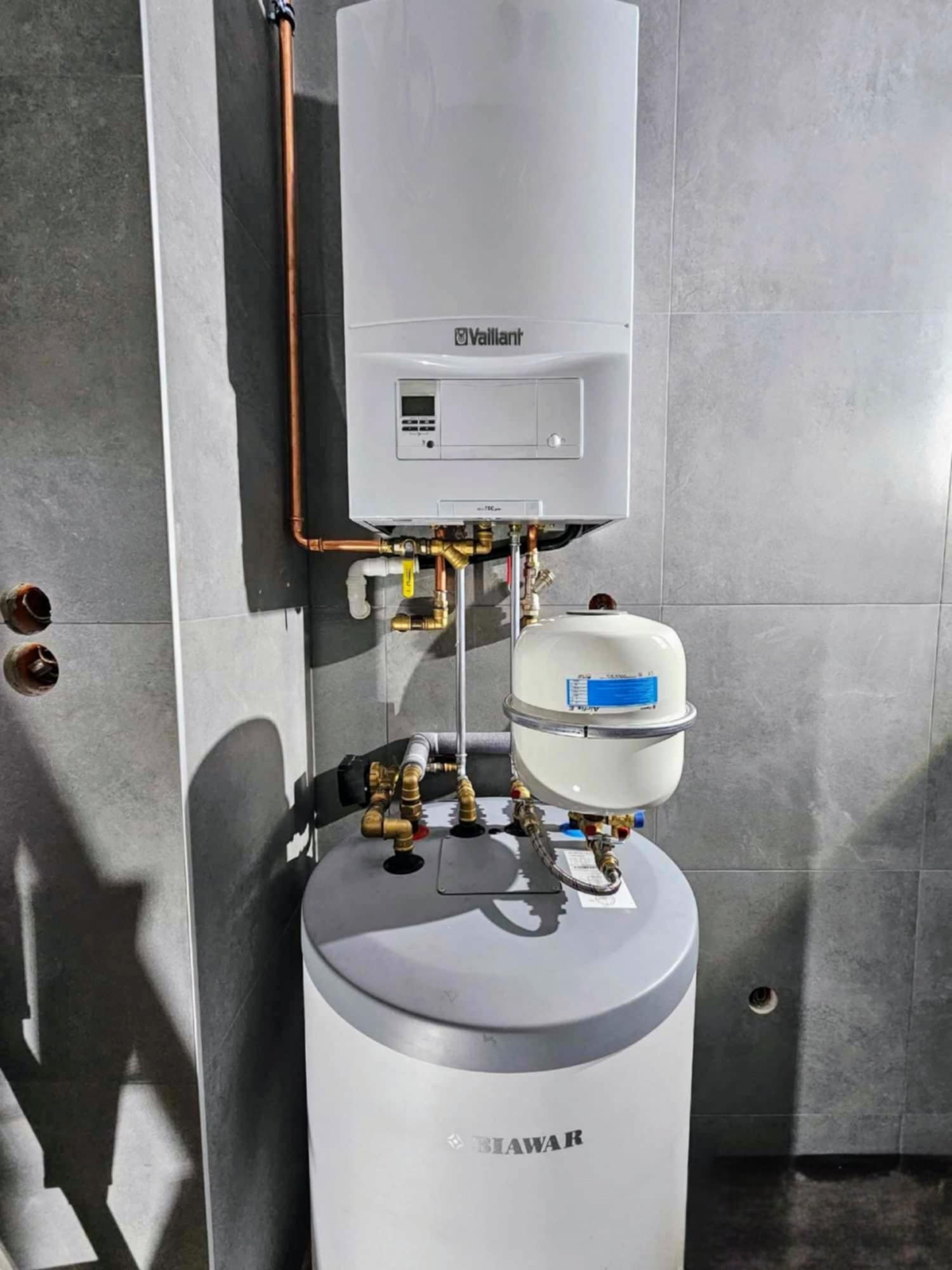 Zamontowany, biały piec gazowy Vaillant z podłączonym zasobnikiem Biawar na tle szarych płytek, widoczne miedziane rury i zawory
