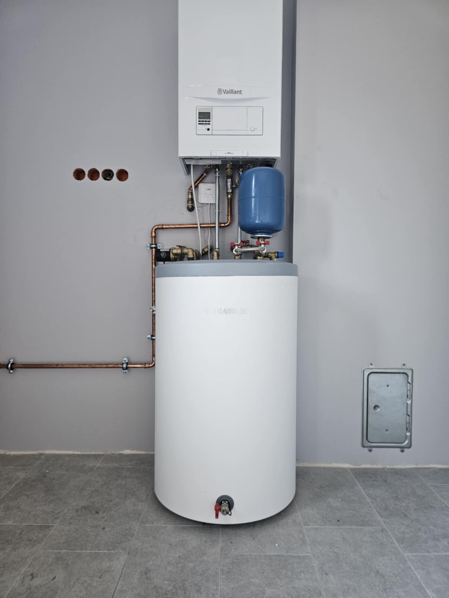 Biały, cylindryczny zasobnik ciepłej wody użytkowej z niebieskim naczyniem wzbiorczym i piecem gazowym Vaillant zamontowane na szarej ścianie z widocznymi miedzianymi rurami i otworami instalacyjnymi.