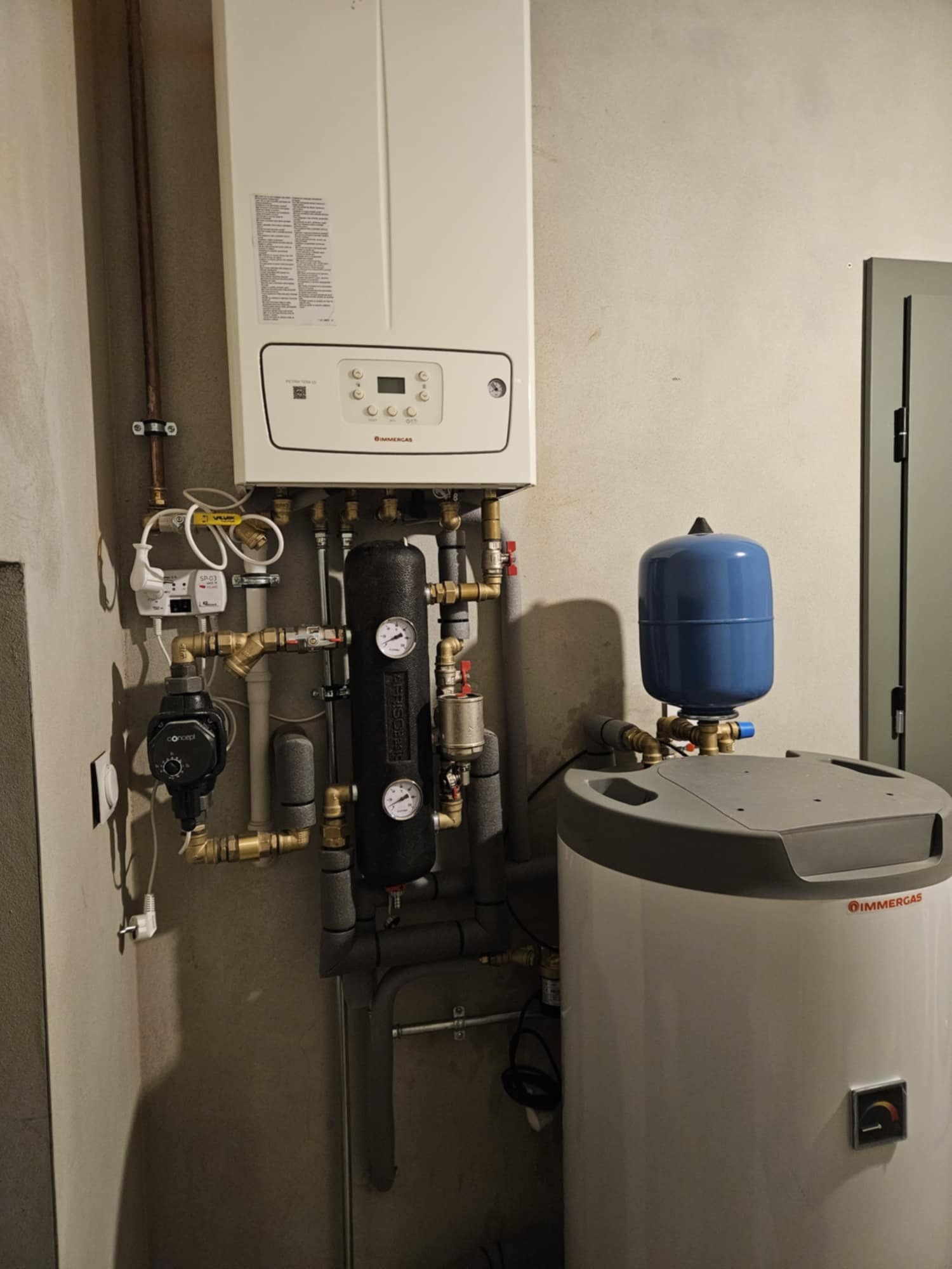 Zestaw kotła grzewczego Immergas z podłączeniami, pompą obiegową Concept i zbiornikiem wyrównawczym, zamontowany na szarej ścianie w pomieszczeniu technicznym.