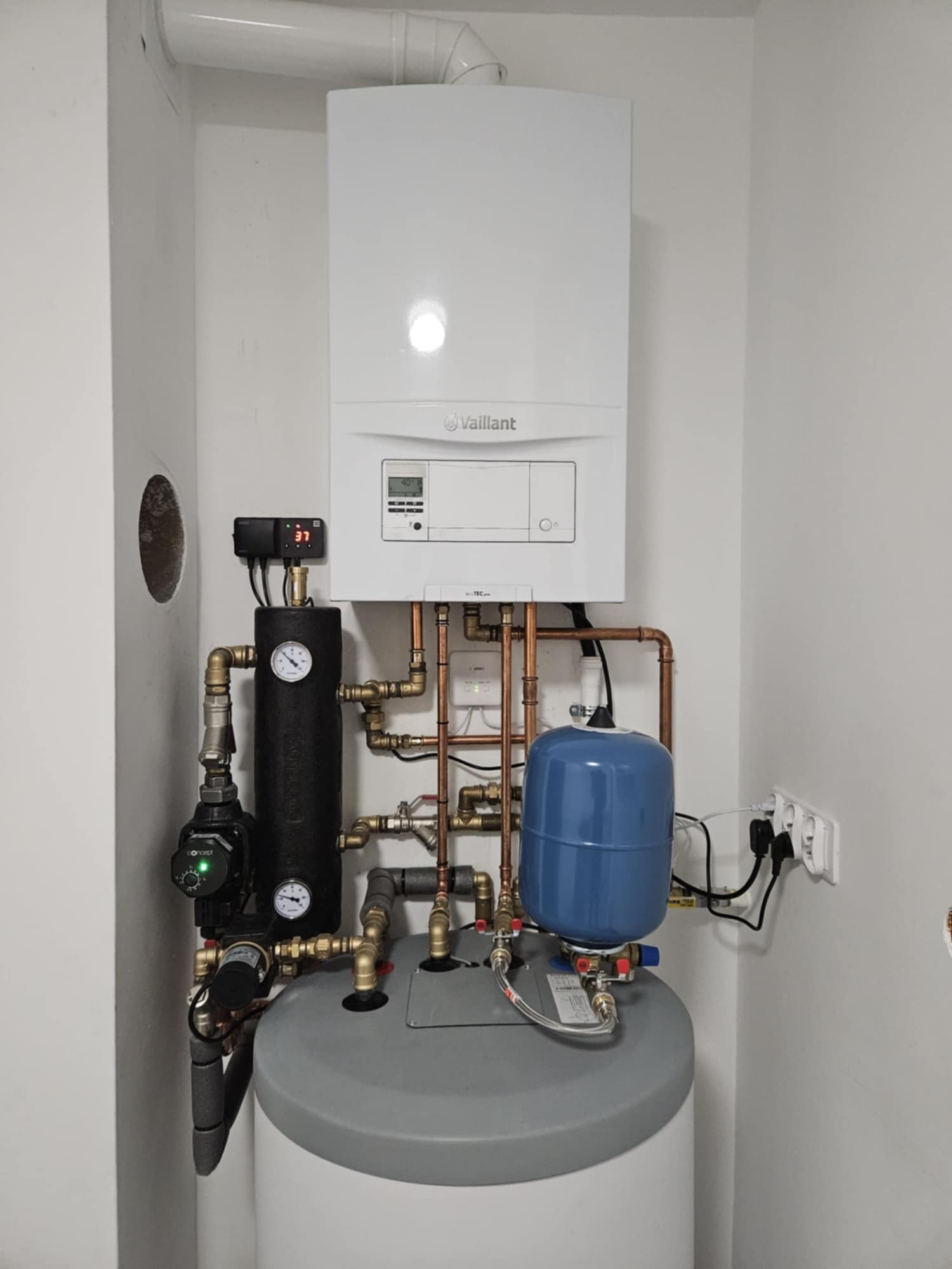 Biały wiszący piec gazowy marki Vaillant z podłączonymi miedzianymi rurami, czarnym zbiornikiem izolowanym i niebieskim zbiornikiem wyrównawczym w białym pomieszczeniu technicznym.