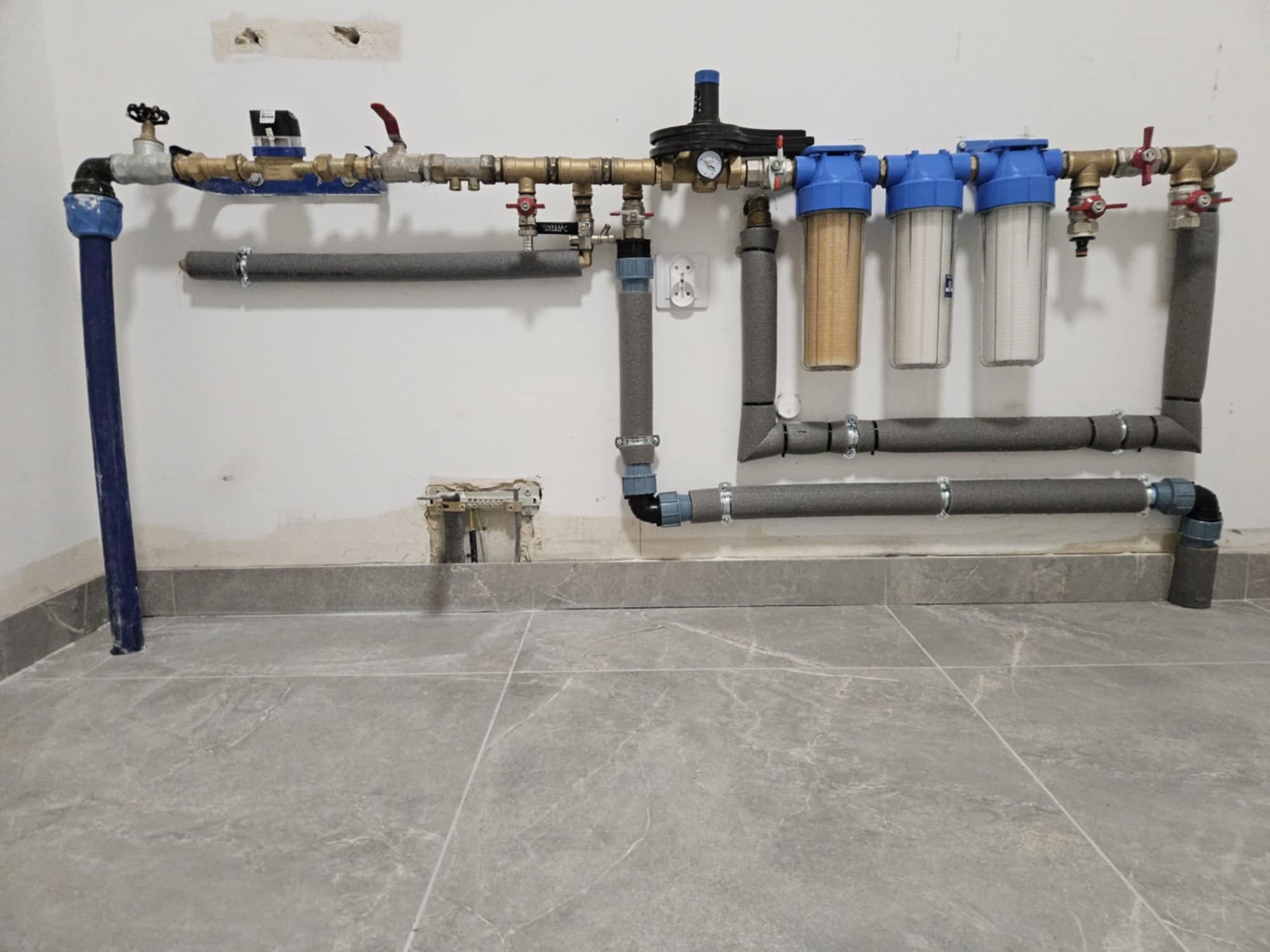 Instalacja wodna z filtrami mechanicznymi i zaworami, rury zaizolowane szarą otuliną, widoczne przyłącze elektryczne obok rur, szare płytki na podłodze, ściana biała z widocznymi śladami po pracach...