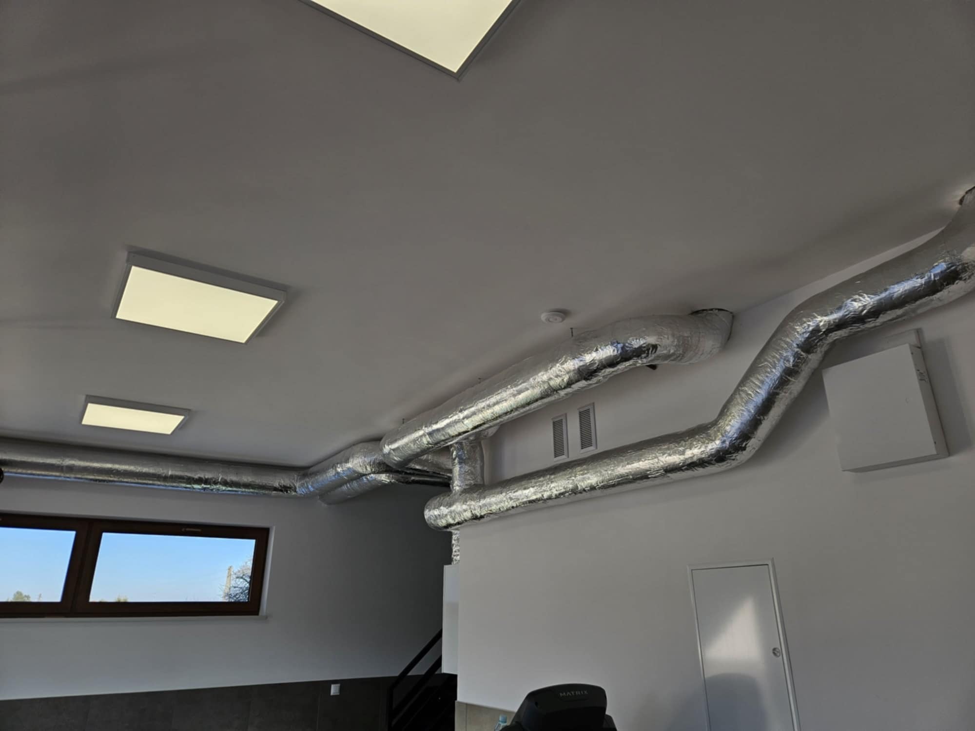 System kanałów wentylacyjnych z folii aluminiowej pod sufitem, widoczne okno i oświetlenie sufitowe, minimalistyczne wnętrze.