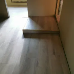 Jasnoszara podłoga z paneli z widocznym progiem wykończeniowym i fragmentem ściany w odcieniach beżu.