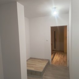 LUX-BUD - Jasne wnętrze po remoncie: białe ściany, drewnopodobne panele podłogowe, schodek z panelami, widok na kolejne pomieszczenie przez otwarte drzwi, nowoczesna lampa sufitowa.