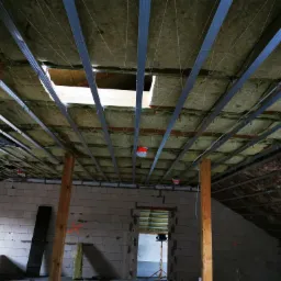 Montaż metalowego stelażu pod płyty gipsowo-kartonowe na poddaszu, widoczna izolacja z wełny mineralnej, surowe ściany z betonu komórkowego, oświetlenie budowlane na statywie.