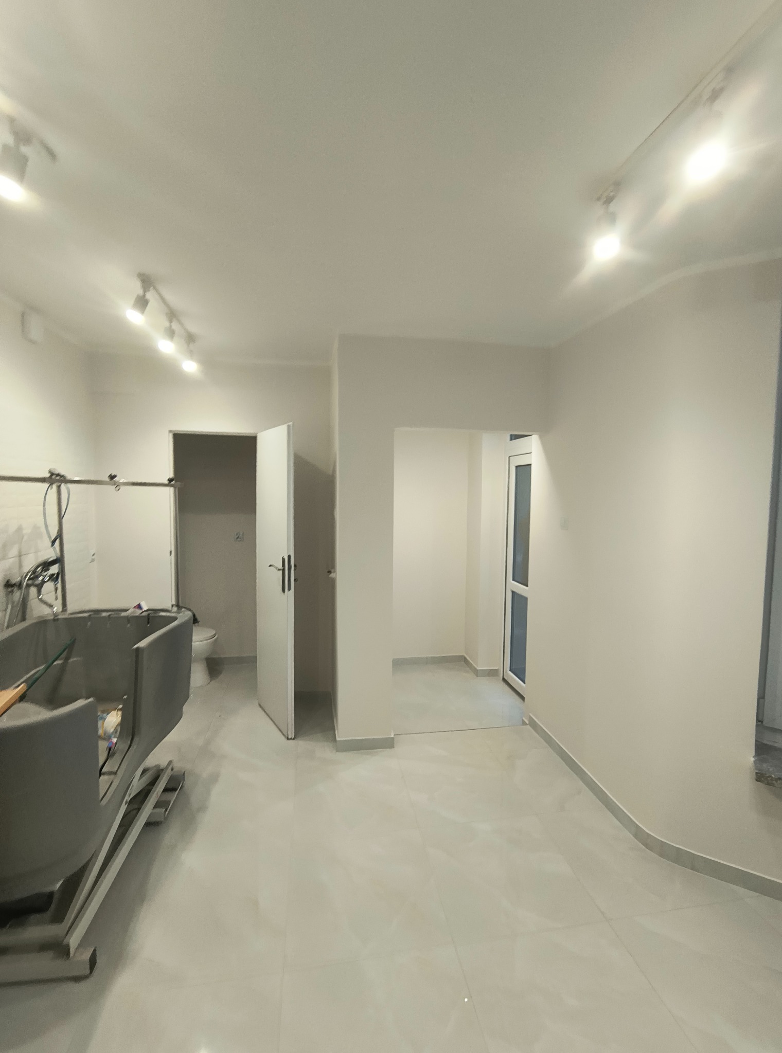 Wnętrze po remoncie: jasne ściany, płytki podłogowe, oświetlenie punktowe. Widoczna wanna w trakcie montażu i otwarte drzwi do łazienki z toaletą.