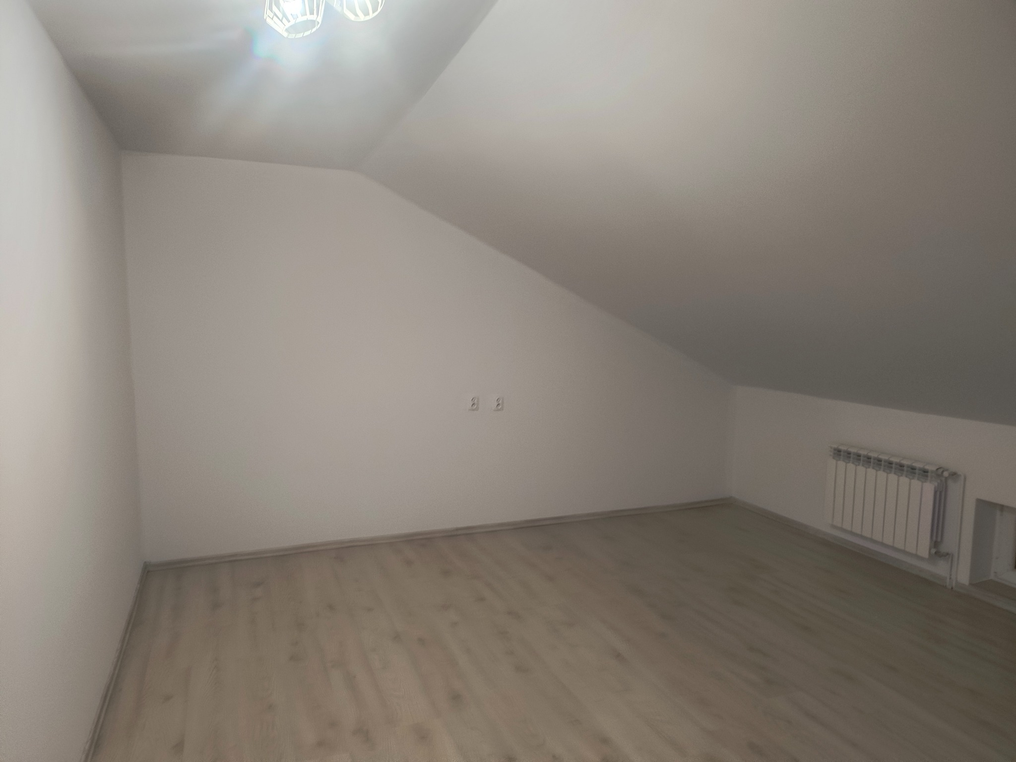 Poddasze po remoncie: jasne panele, białe ściany, grzejnik. Widoczne gniazdka elektryczne. Ukośne ściany charakterystyczne dla poddasza.