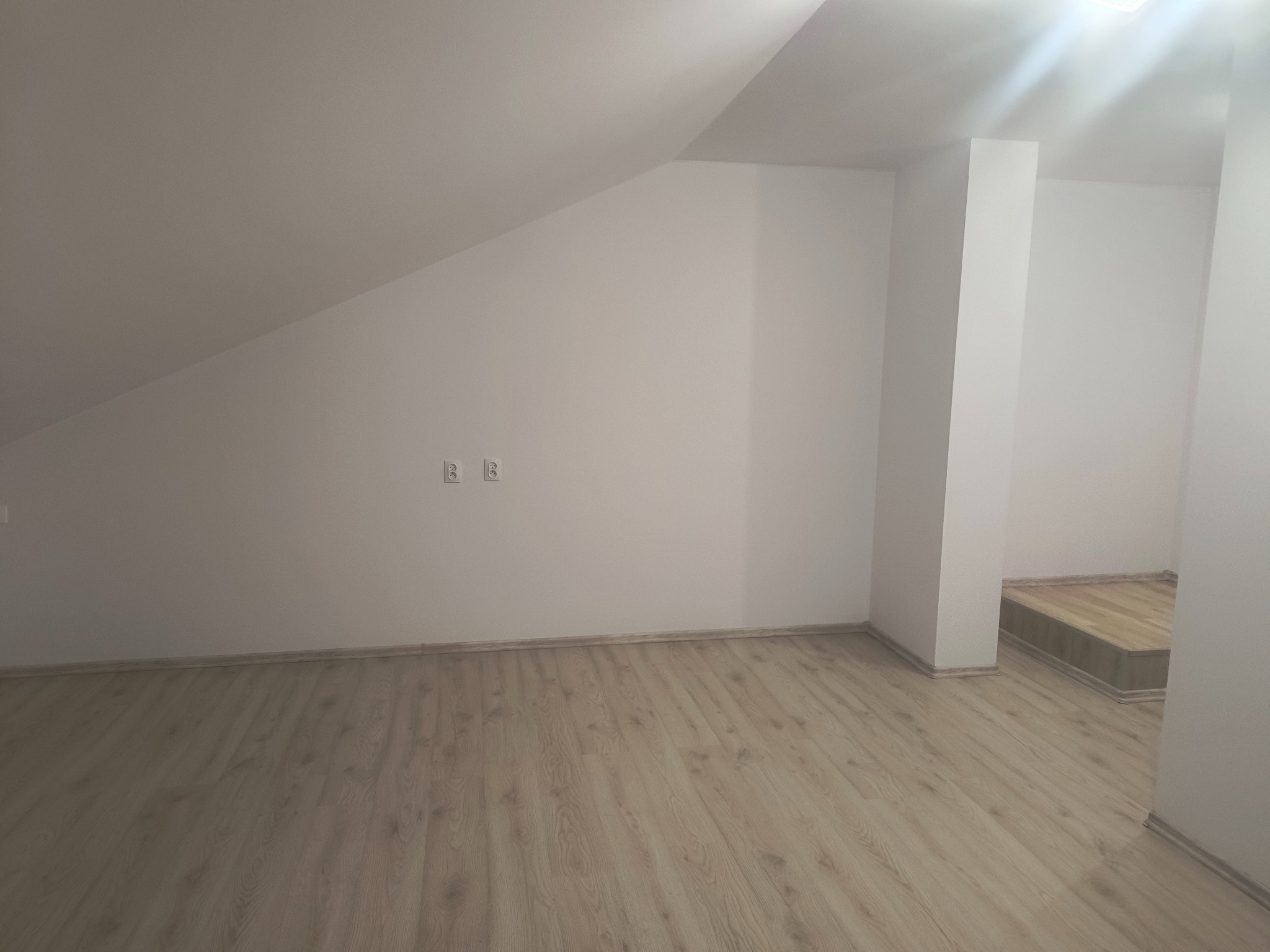 Jasne, odnowione poddasze z panelami podłogowymi i świeżo pomalowanymi ścianami. Widoczne gniazdka elektryczne i fragment schodów w przejściu.