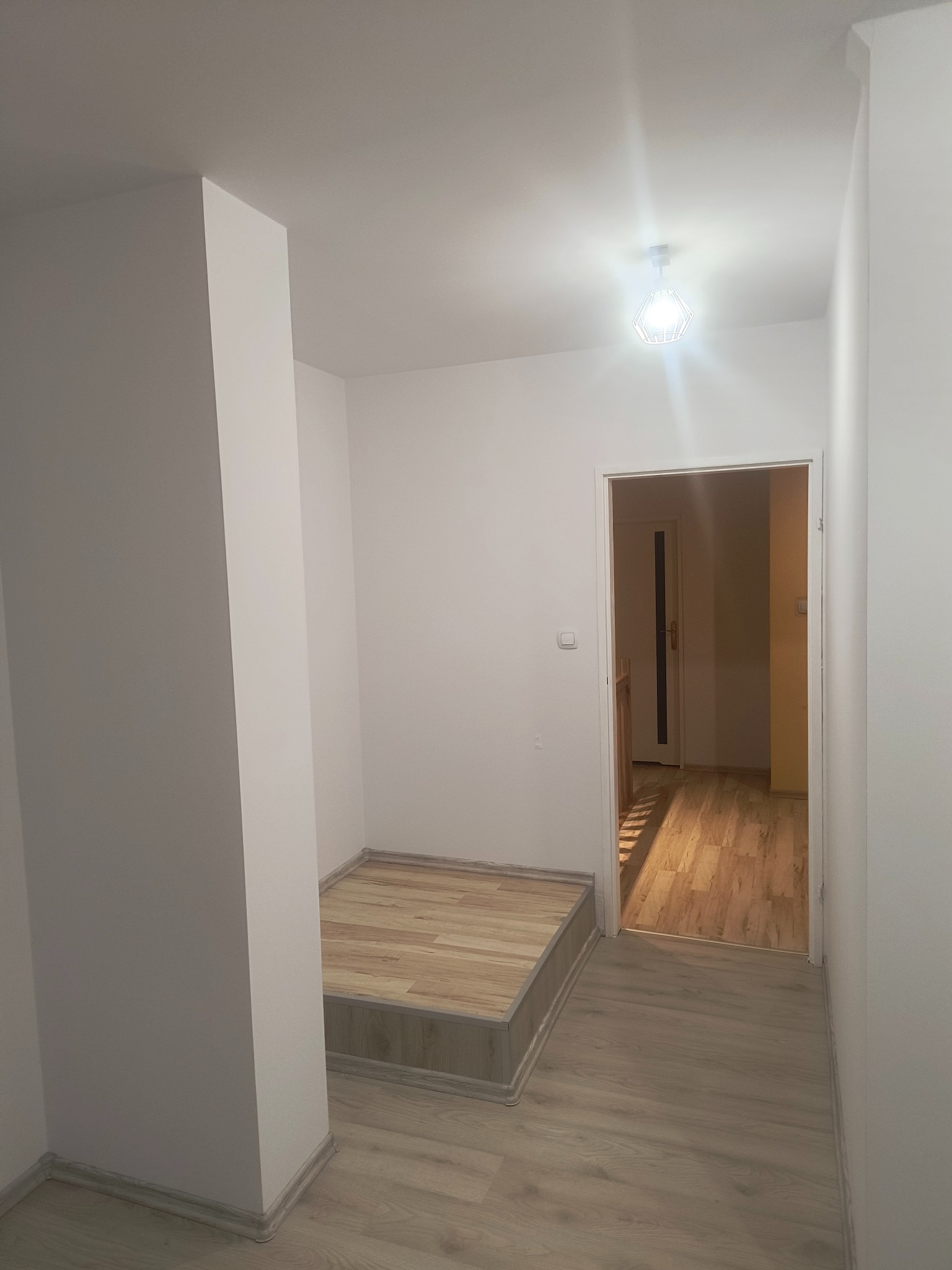 Jasne wnętrze po remoncie: białe ściany, drewnopodobne panele podłogowe, schodek z panelami, widok na kolejne pomieszczenie przez otwarte drzwi, nowoczesna lampa sufitowa.