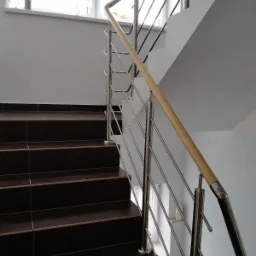 Schody z ciemnymi płytkami i balustradą ze stali nierdzewnej z drewnianym poręczą, widok z dołu na górę.