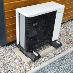 Biała pompa ciepła Hewalex zamontowana na betonowych bloczkach obok drewnianego budynku, otoczona drobnymi kamieniami.