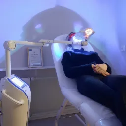 Pacjentka poddawana zabiegowi z użyciem specjalistycznego urządzenia emitującego światło w gabinecie medycyny estetycznej, widoczny certyfikat na ścianie.