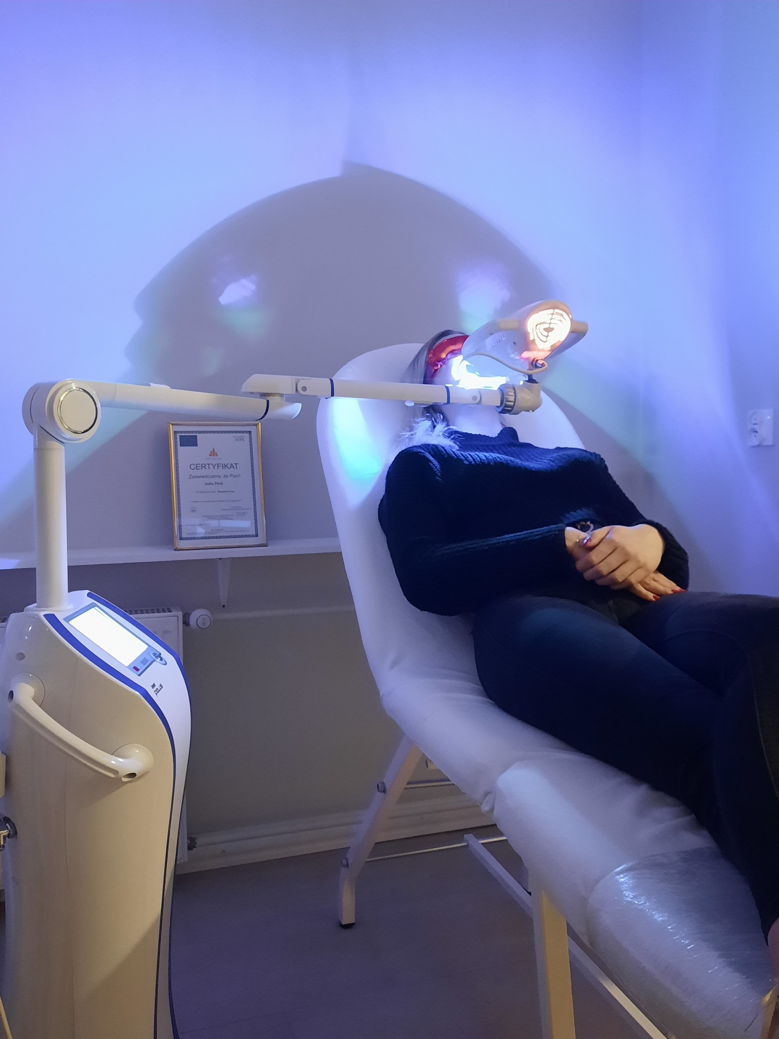Pacjentka poddawana zabiegowi z użyciem specjalistycznego urządzenia emitującego światło w gabinecie medycyny estetycznej, widoczny certyfikat na ścianie.