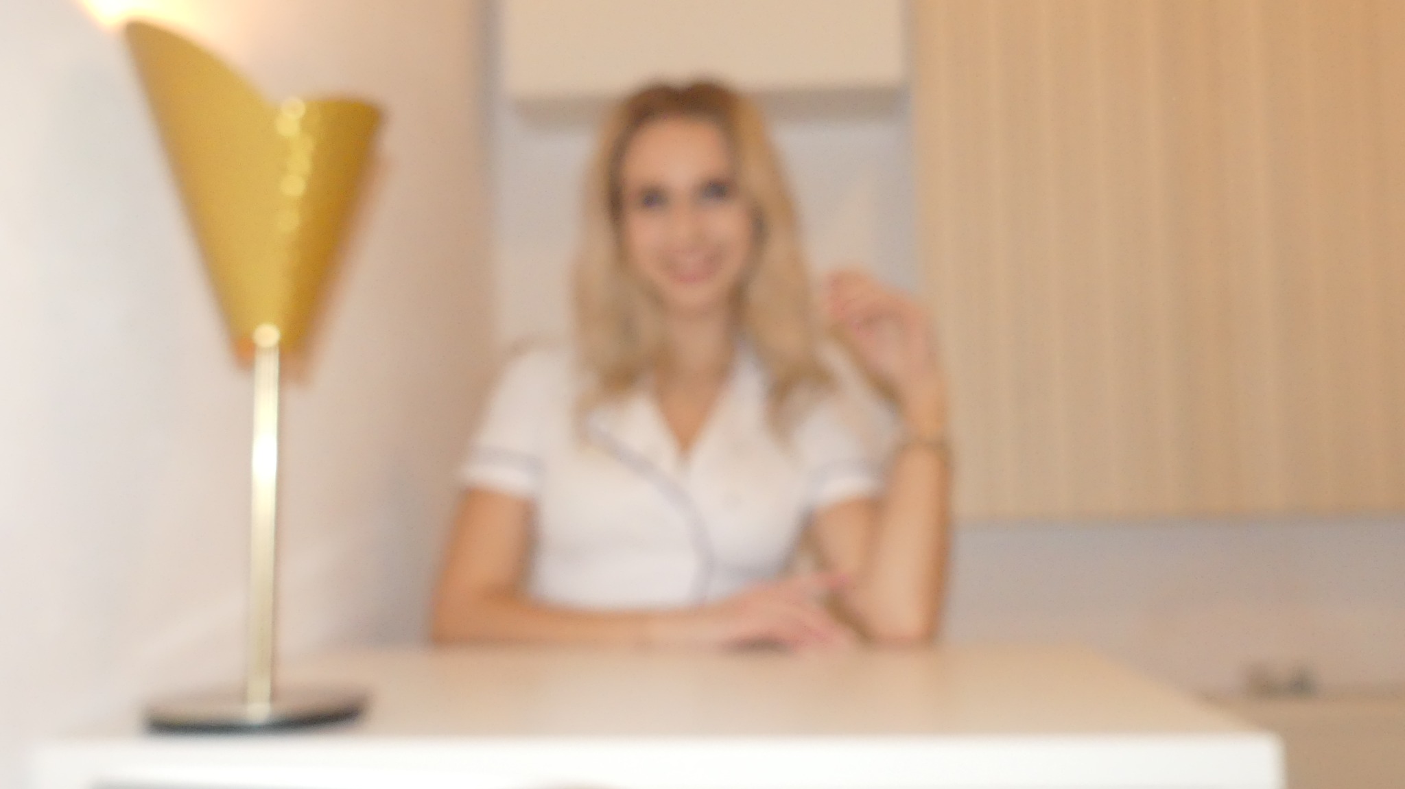 Portret uśmiechniętej blondynki w białym uniformie medycznym siedzącej za białym stołem, z dekoracyjną złotą lampą po lewej stronie kadru.