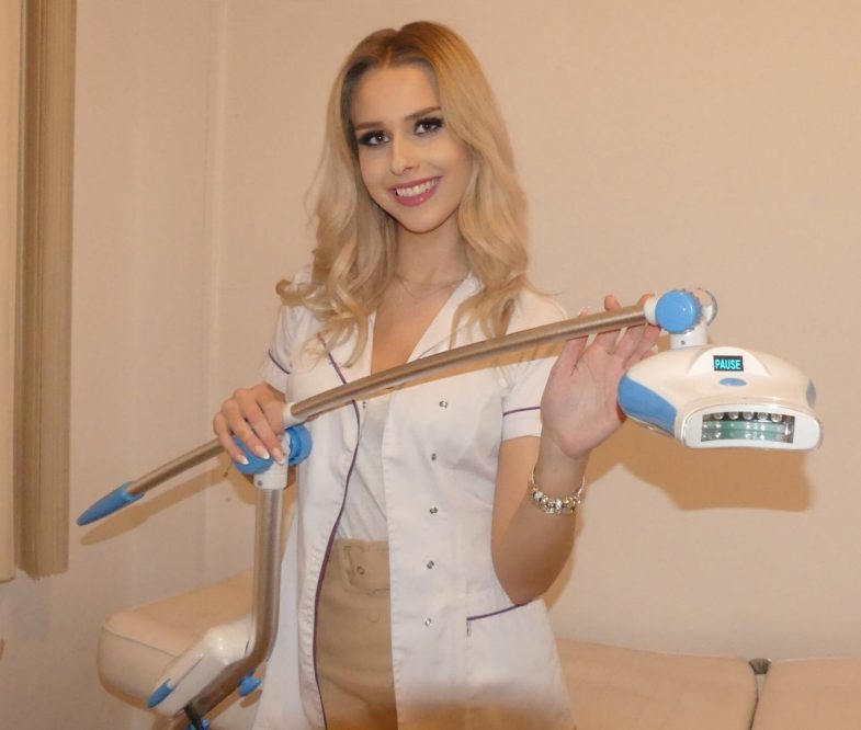 Uśmiechnięta blondynka w białym uniformie prezentuje lampę do wybielania zębów z napisem 'PAUSE' na wyświetlaczu.