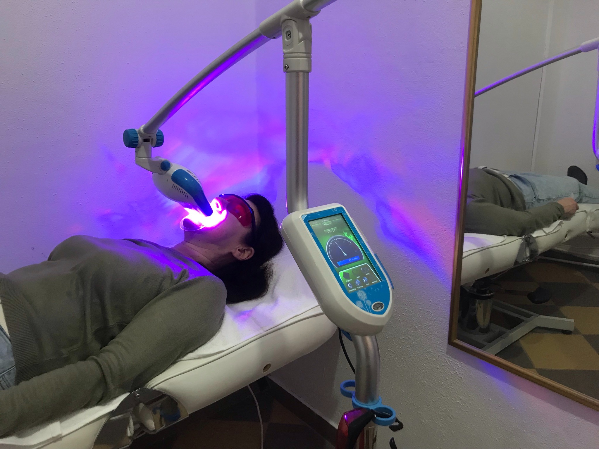 Pacjentka leży na fotelu zabiegowym podczas wybielania zębów za pomocą lampy UV, widoczne okulary ochronne i panel sterowania urządzenia.