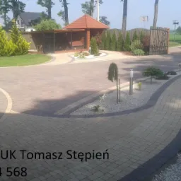 Ułożona kostka brukowa w odcieniach beżu i szarości tworząca elegancki podjazd z dekoracyjnymi obrzeżami, prowadzący do bramy wjazdowej i altany ogrodowej na posesji.