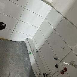 Białe płytki ścienne w trakcie montażu w łazience, widoczne narzędzia glazurnicze i elementy instalacji. Surowy beton na podłodze. Ujęcie z góry, ukośne.