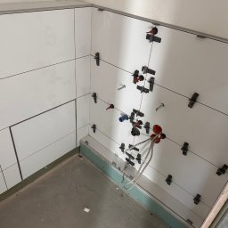 Białe, błyszczące płytki ścienne w trakcie montażu, z użyciem klipsów poziomujących. Widoczne wystające rury instalacji wodnej i fragment podłogi w surowym stanie.