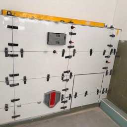 Usługi Ogólnobudowlane Michał Lesznik - Białe płytki ścienne w trakcie montażu, z użyciem klipsów poziomujących i poziomicy, widoczne elementy instalacji wodnej w surowej łazience.