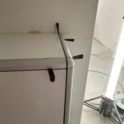 Fragment białej ściany z widocznymi śladami po szpachlowaniu i mocowaniach, oświetlonej lampą budowlaną. Detal wykończenia wnętrza w trakcie prac.