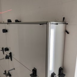 Białe płytki w rogu łazienki, wyrównane za pomocą klipsów i lasera, ukazujące precyzję montażu. Widoczne detale wykończenia i narzędzia glazurnicze.